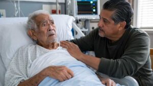 Hombre acompañando y consolando a un adulto mayor en una cama de hospital